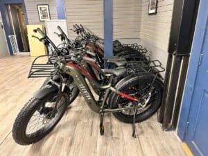 A line of Aventon electric bikes is displayed in a store. Each bike features a black frame, a front cargo rack, and fat tires. Perfect for exploring destinations like Pikes Peak RV Park or Gulf Beach RV Resort, they are neatly arranged side by side on a wooden floor with a striped back wall.