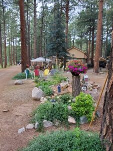 A serene outdoor setting featuring a lush Memorial Garden with various plants and small decorative houses. A cluster of colorful chairs sits under a canopy near a beige house, surrounded by tall pine trees. A hanging flower basket is prominently displayed in the foreground.