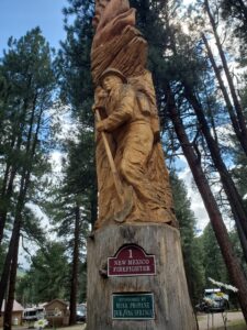 Wooden statue of a firefighter holding a shovel, carved into a tree trunk. The sign below reads "New Mexico Firefighter" followed by "Sponsored by Mesa Propane Dur/Pag Springs." Tall trees and a partly cloudy sky are visible in the background. A highlight of the Wood Carvings Tour at Junction West Vallecito Resort.