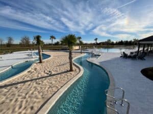 A serene outdoor pool area with a winding lazy river, palm trees, lounge chairs, and clear blue skies. The poolside area at the Tiger's Trail RV Resort is spacious and has several seating areas with tables and chairs. The landscape in the background includes fields and trees.