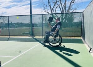 On a sunny outdoor court, a wheelchair user swings a paddle in an exhilarating pickleball game, perhaps under the guidance of expert Pickleball Coaching. The court, encircled by a chain-link fence, is graced by a tree and blue sky in the backdrop.